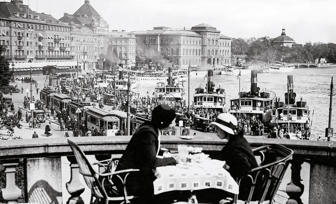 Historisk bild från Stockholm. Två kvinnor sitter på en uteservering. I bakgrunden syns Grand Hotel och vatten