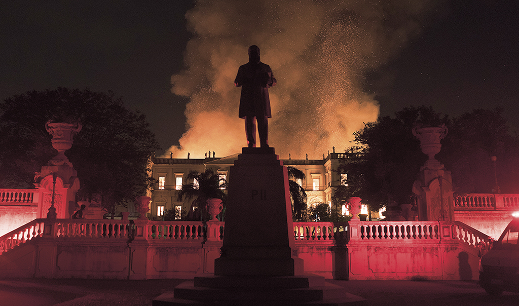 En stor brand slukar samlingarna bestående av 20 miljoner föremål i det 200 år gamla National­museet i Rio de Janeiro. Himlen är rödtonad och i förgrunden syns en staty.