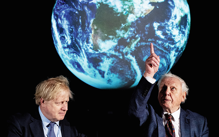 Boris Johnson och David Attenborough snett framför en bild på jorden. David Attenborough blickar uppåt och pekar.