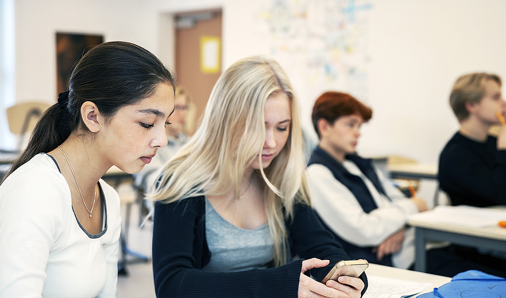 Två unga kvinnliga elever sitter vid en skolbänk i ett klassrum och tittar koncentrerat på en mobilskärm.