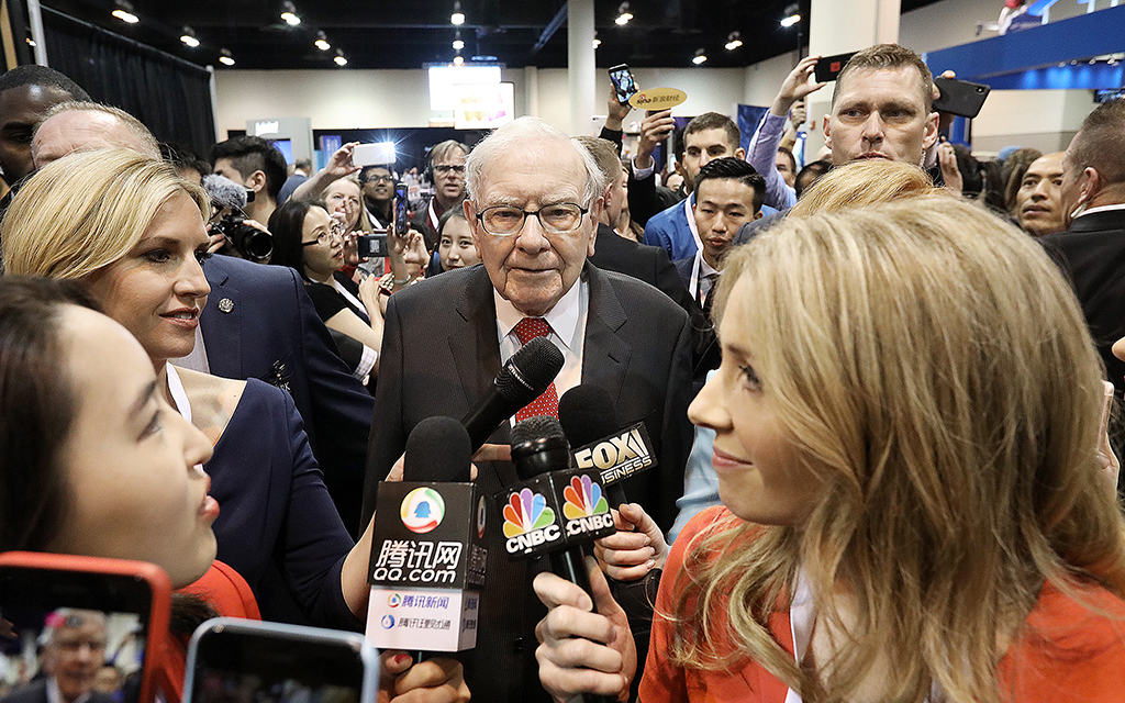 Warren Buffett omgiven av ett stort antal journalister som håller upp mikrofoner framför honom.