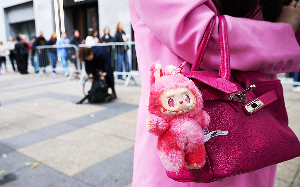 En rosaklädd kvinna Med en rosa väska på armen. På väskan hänger en rosa Labubufigur och i bakgrunden syns kön till den affär i Berlin där den är köpt.