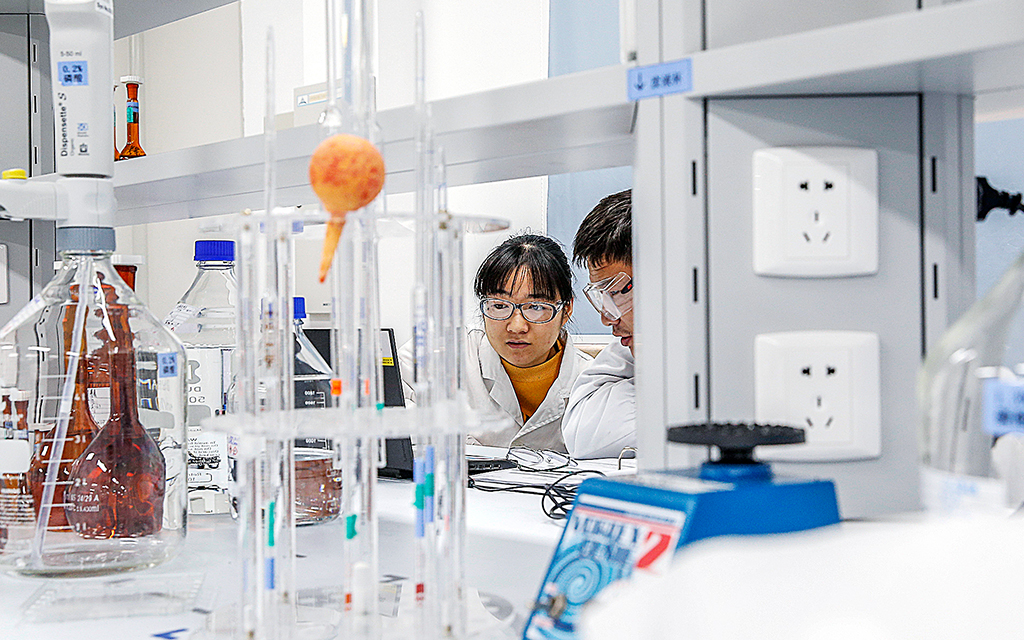 En kvinna och en man i labbkläder sitter och tittar på något på en dator i en labbmiljö. I förgrunden syns glasflaskor, burkar och annat laboratoriematerial