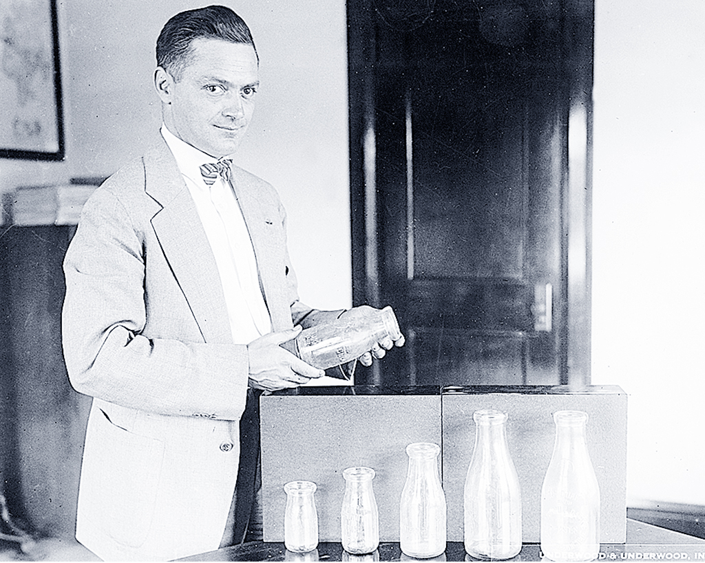 En man i kavaj och fluga står bredvid ett bord och visar upp standardiserade mjölkflaskor för kameran. 1920-tal.