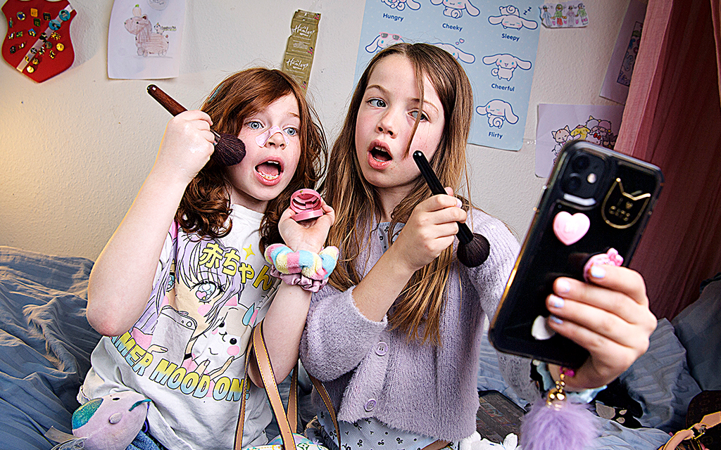 Två unga flickor sminkar sig och gör miner in i en mobiltelefon.