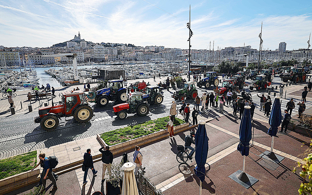 En lång rad röda pch blå traktorer passerar åskådare som står och tittar på dem på en solig hamnpromenad i Marseille. I bakgrunden syns vatten och stora båtar.