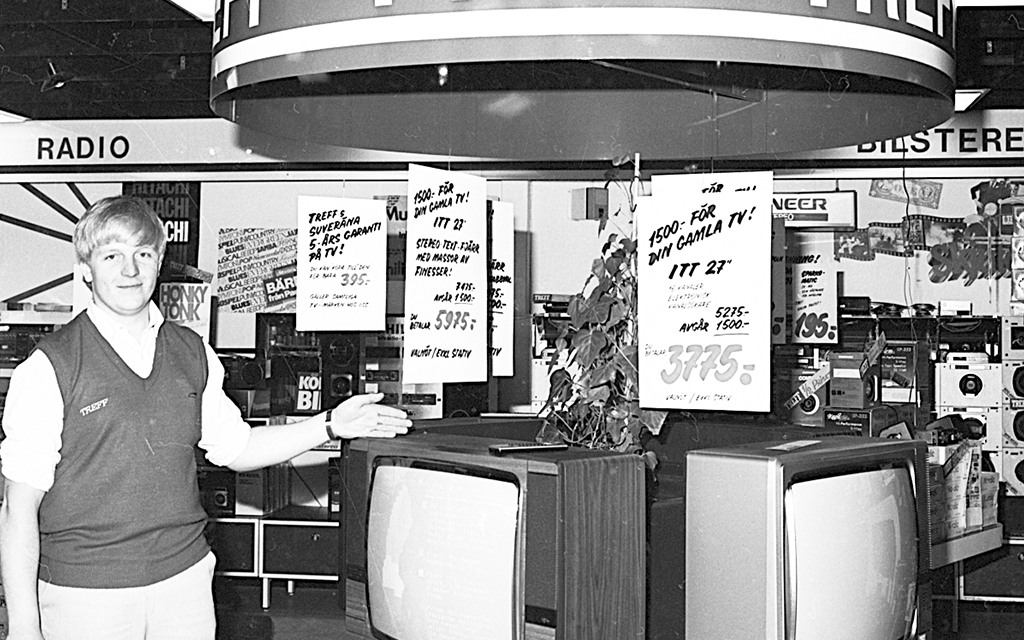 Svartvit bild från 1970. En butiksanställd står bredvid några tv-apparater och pekar på dem. Ovanför hänger stora prisskyltar.