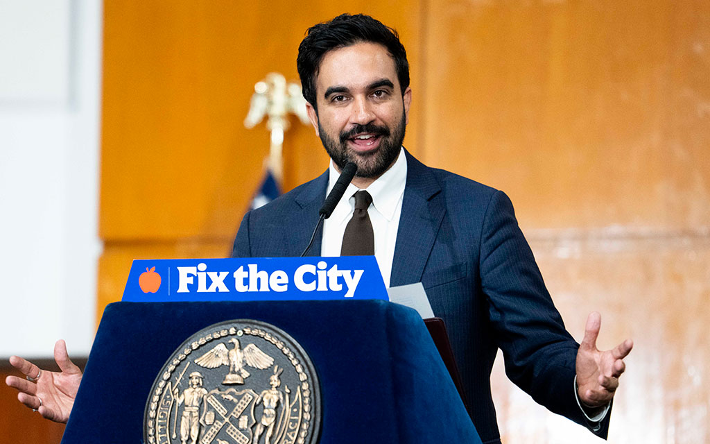 Man i kostym talar vid mikrofon med en skyltdocka framför sig som visar 'Fix the City'.
